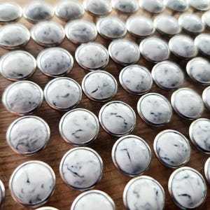 May include: A close-up of a collection of white marble buttons with silver metal rims. The buttons are arranged in a grid pattern on a wooden surface.