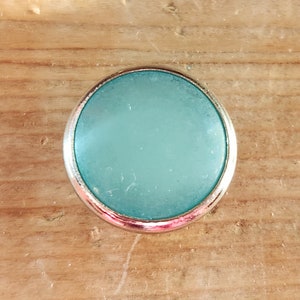 Puede incluir: Un cabuj&oacute;n redondo de color azul claro con un engaste plateado. El cabuj&oacute;n es liso y tiene un acabado ligeramente esmerilado.