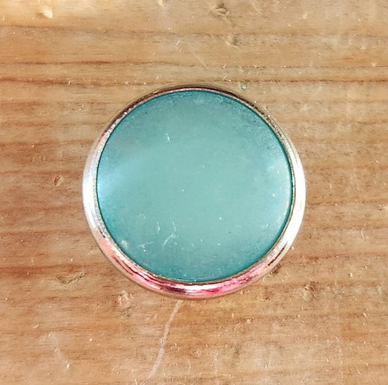 Puede incluir: Un cabuj&oacute;n redondo de color azul claro con un engaste plateado. El cabuj&oacute;n es liso y tiene un acabado ligeramente esmerilado.