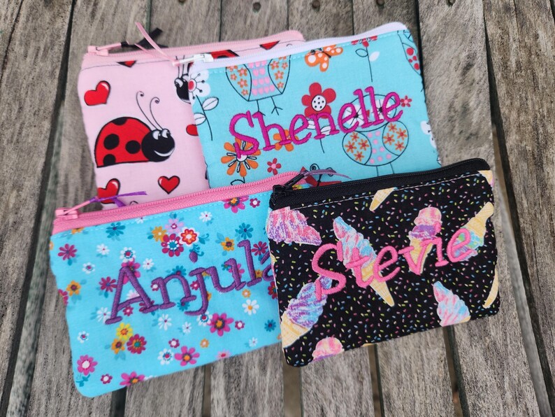 May include: Four rectangular zippered pouches featuring various fabric patterns. One is pink with ladybugs and hearts, one is light blue with floral bird motifs and the name Shenelle, one is light blue with flowers and the name Anjula, and one is black with ice cream cones and the name Stevie.