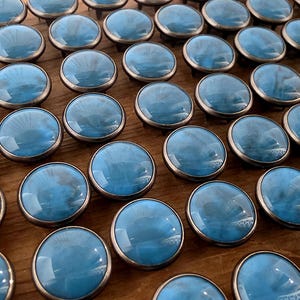 May include: A collection of round furniture knobs featuring a smooth, glossy light blue surface with a subtle marbled effect, encased in a metallic silver-toned rim. Each knob has a domed top and a circular base, arranged in a grid pattern.