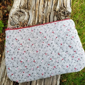K&ouml;nnte beinhalten: Eine graue gesteppte Rei&szlig;verschlusstasche mit Blumenmuster und einem burgunderfarbenen Rei&szlig;verschluss. Die Tasche liegt auf einem St&uuml;ck verwittertem Holz.