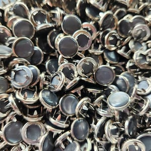 May include: A large pile of silver and black metal buttons with a pointed prong on the back. The buttons have a round, flat top with a shiny, iridescent finish.