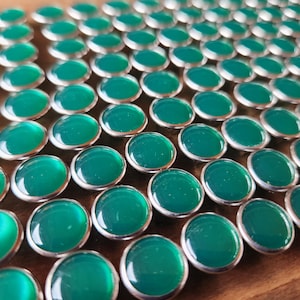 May include: A collection of oval-shaped, polished green gemstones set in silver-colored metal bezels. The gemstones are arranged in rows, creating a pattern. The background is a warm, wooden surface.