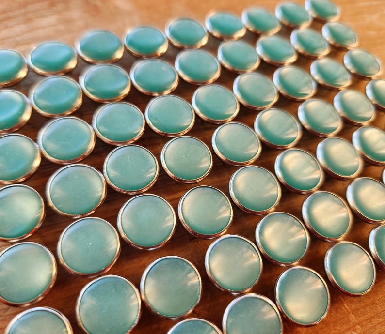 Puede incluir: Primer plano de una gran cantidad de botones redondos de color azul turquesa con bordes plateados. Los botones est&aacute;n dispuestos en una cuadr&iacute;cula sobre una superficie de madera.
