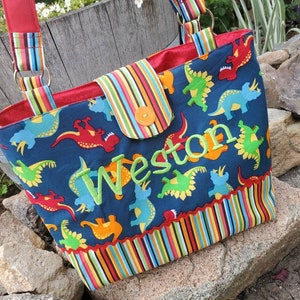 May include: A blue fabric tote bag with a colourful striped border and a dinosaur print. The bag has a red lining and a flap with a button closure. The name "Weston" is embroidered on the front of the bag in green.