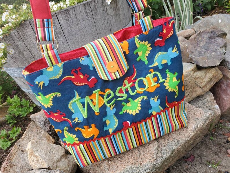 May include: A blue fabric tote bag with a colourful striped border and a dinosaur print. The bag has a red lining and a flap with a button closure. The name "Weston" is embroidered on the front of the bag in green.
