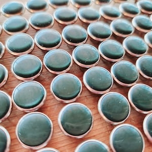 May include: A collection of round, dark green cabochons with silver-colored metal rims. The polished stones have a smooth, reflective surface. The cabochons are arranged on a light brown wooden surface.