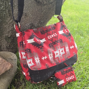May include: A red and black crossbody bag featuring a geometric pattern with white accents, a black flap, and a matching small rectangular pouch attached to the front. The bag includes a black adjustable strap connected by silver-toned metal rings.