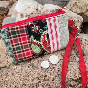 May include: A small, patchwork coin purse with a red zipper and wrist strap. The purse features various fabric patterns, including plaid, paisley, and floral designs. Two coins are placed near the purse.