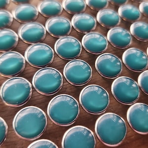 May include: A collection of teal cabochon buttons with a silver-colored metal rim. The buttons have a smooth, glossy surface and are arranged in a diagonal pattern. The teal color is a vibrant, eye-catching hue. These buttons are ideal for sewing and craft projects.
