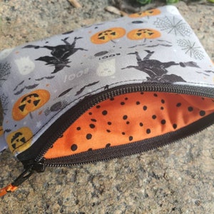May include: A small grey zippered pouch with a Halloween theme. The pouch has a black zipper and a black and orange polka dot lining. The exterior of the pouch features a repeating pattern of bats, ghosts, pumpkins, and spiderwebs.