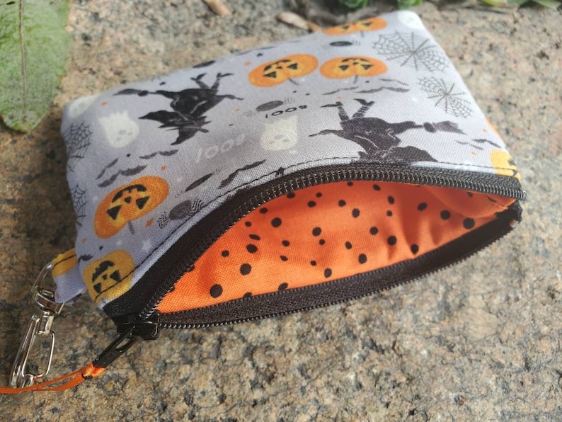 May include: A small grey zippered pouch with a Halloween theme. The pouch has a black zipper and a black and orange polka dot lining. The exterior of the pouch features a repeating pattern of bats, ghosts, pumpkins, and spiderwebs.