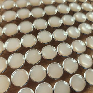 May include: A close-up view of a large quantity of white pearl-like buttons with silver metal rims. The buttons are arranged in rows on a wooden surface.