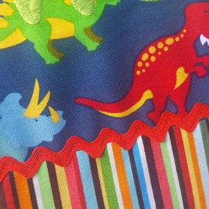 May include: Blue fabric with a colourful striped border. The fabric features a repeating pattern of dinosaurs in various colours, including green, red, blue, and yellow.