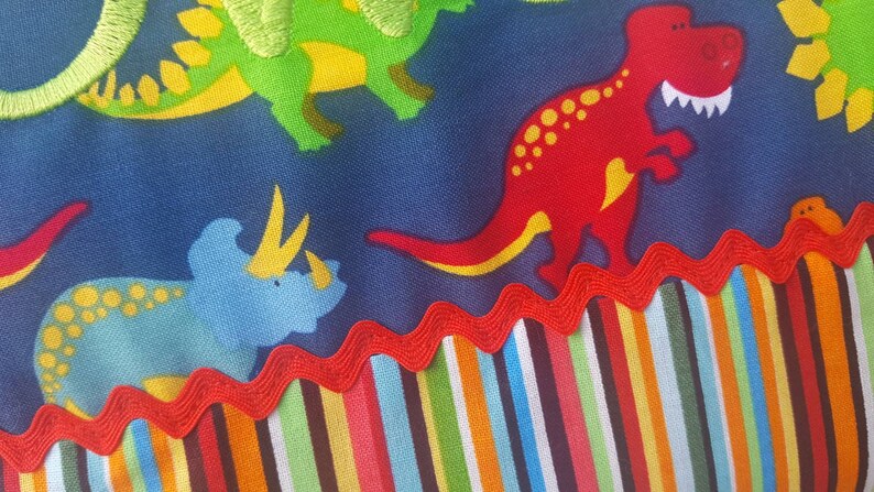 May include: Blue fabric with a colourful striped border. The fabric features a repeating pattern of dinosaurs in various colours, including green, red, blue, and yellow.