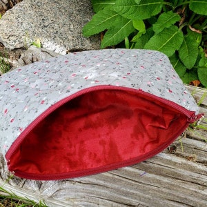 K&ouml;nnte beinhalten: Eine graue, gesteppte Rei&szlig;verschlusstasche mit rotem Blumendruck und rotem Futter. Die Tasche ist offen und liegt auf einer Holzoberfl&auml;che.