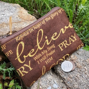 May include: A brown zippered pouch with a yellow and brown floral pattern. The pouch has the words "Rejoice in hope of the glory of" and "believe" printed on it. The pouch is sitting on a rock with some coins next to it.