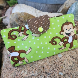 May include: A green wallet with a brown polka-dot flap and a yellow button closure. The wallet features a repeating pattern of cartoon monkeys with brown fur and white faces. The background is a light green with white polka dots.