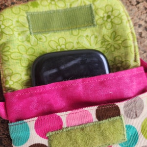 May include: A pink and green fabric pouch with a polka dot pattern. The pouch has a flap that closes with a velcro strap. A black rectangular object is inside the pouch.