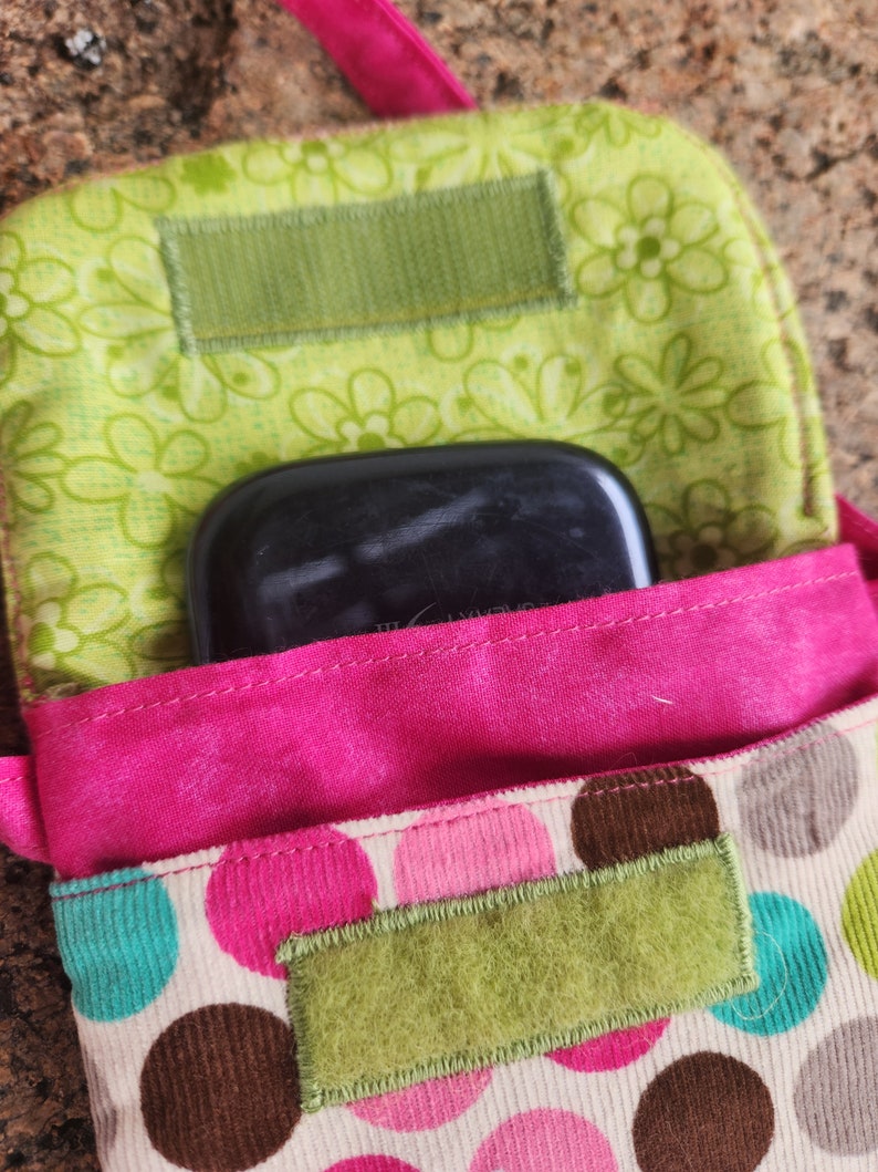 May include: A pink and green fabric pouch with a polka dot pattern. The pouch has a flap that closes with a velcro strap. A black rectangular object is inside the pouch.