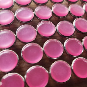 May include: A close-up of a large quantity of pink glass cabochons with silver settings. The cabochons are arranged in rows on a wooden surface.