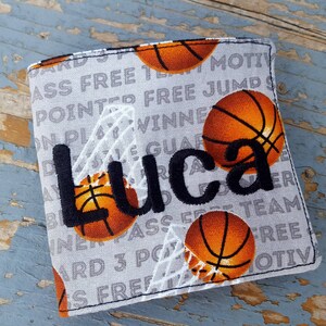 May include: A gray fabric wallet with a basketball theme. The wallet has a white basketball net design and multiple basketballs in orange and black. The name "Luca" is embroidered in black on the front of the wallet.