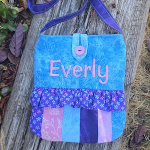 May include: A fabric crossbody bag featuring a blue dragonfly print top section with the name Everly embroidered in pink. It has a purple ruffled trim, a vertical striped lower section in shades of pink and blue, and a purple strap with a button.