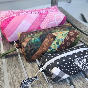 May include: Three zippered pouches made of fabric with different patterns. The top pouch is pink with pink and white stripes. The middle pouch is brown with green, brown, and white stripes. The bottom pouch is black with white polka dots and a black and white checkered pattern.