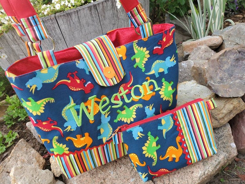 May include: A blue and red tote bag with a colourful striped trim and a dinosaur print. The bag has a zipped pocket and is personalised with the name "Weston".