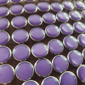 May include: A close-up of a large quantity of round, purple buttons with silver metal rims. The buttons are arranged in rows and are all the same size.