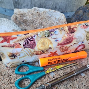 May include: A zippered pencil case with a seashell pattern. The case is made of fabric and has an orange zipper. The case is lying on a rock with a highlighter, scissors, and a pen next to it.
