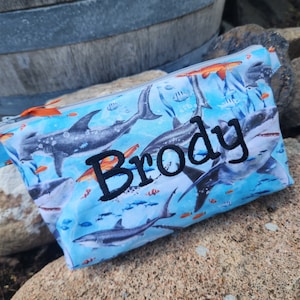 May include: A blue zippered pouch with a shark and fish print. The pouch has the name "Brody" embroidered on it in black thread.
