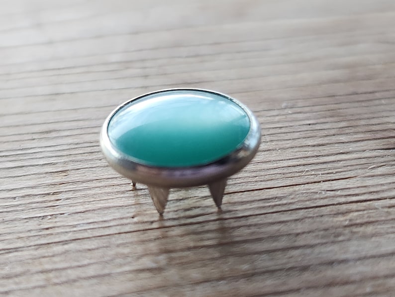 Puede incluir: Un cabuj&oacute;n de vidrio azul turquesa engastado en una base de metal plateado con tres puntas. El cabuj&oacute;n es liso y brillante.