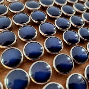 May include: A collection of navy blue buttons with silver-colored rims. The buttons are arranged in a grid pattern, with a glossy finish. The buttons are round and have a slightly domed shape, suggesting they are for clothing or crafts.