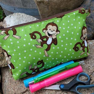 May include: Green zippered pouch with a white polka dot pattern and brown monkeys with white eyes. The pouch is lying on a pile of rocks with three pens and a pair of scissors.