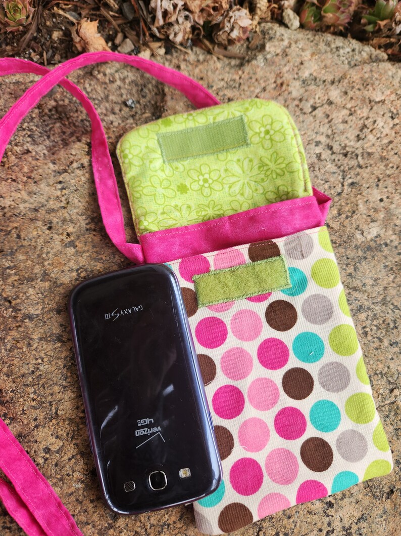 May include: A pink and green fabric phone case with a polka dot pattern and a flap closure. The case has a strap for carrying over the shoulder. A black Samsung Galaxy S III phone is inside the case.
