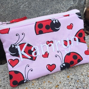 May include: A light pink zippered pouch featuring a repeating pattern of red ladybugs with black spots and small red hearts. The name Kamryn is embroidered in white script across the center of the pouch, which has a matching light pink zipper.