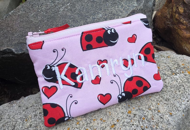 May include: A light pink zippered pouch featuring a repeating pattern of red ladybugs with black spots and small red hearts. The name Kamryn is embroidered in white script across the center of the pouch, which has a matching light pink zipper.