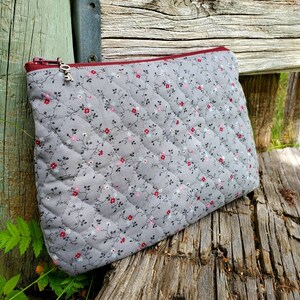 K&ouml;nnte beinhalten: Eine graue, gesteppte Rei&szlig;verschlusstasche mit Blumenmuster und einem silbernen Rei&szlig;verschluss. Die Tasche liegt auf einer Holzoberfl&auml;che.