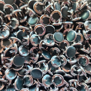 May include: A close-up view of a pile of silver-toned metal rivets with green plastic tops. The rivets have a star-shaped base and are designed for attaching fabric or leather.