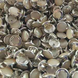 May include: A close-up of a pile of silver and off-white round buttons with a pointed metal base. The buttons are used for attaching to fabric.