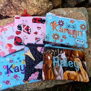 May include: A collection of five rectangular fabric zipper pouches featuring various patterns including ladybugs, flowers, owls, ice cream cones, and horses. Each pouch is personalized with an embroidered name in a distinct thread color.