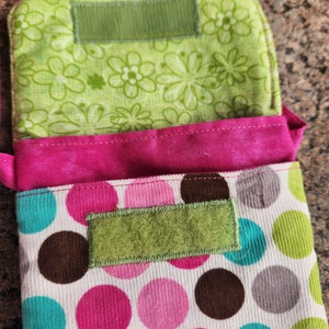 May include: A pink and white fabric bag with brown, pink, and turquoise polka dots. The bag has a green fabric flap with a velcro closure.
