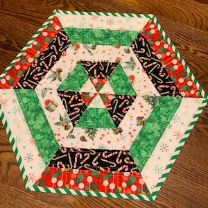 May include: A hexagonal quilted table mat featuring a festive holiday pattern. The design includes alternating bands of white fabric with pinecones and candy canes, solid green fabric, and red fabric with lollipops, finished with a green and white striped border.