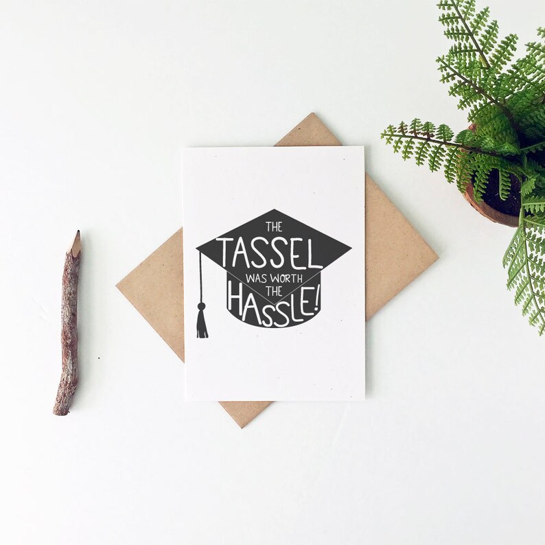 May include: A graduation card with a black mortarboard design and the text "THE TASSEL WAS WORTH THE HASSLE!" is displayed on a white background. The card is placed on a brown envelope. A pencil and a potted fern are also in the image.