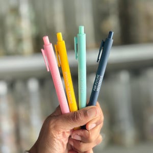 Op de afbeelding: Een hand houdt vier kleurrijke pennen vast: roze, geel, blauwgroen en marineblauw. Elke pen heeft een andere witte tekst: "PER MY LAST EMAIL", "THANKS IN ADVANCE", "PLEASE ADVISE" en "FOR FUTURE REFERENCE".