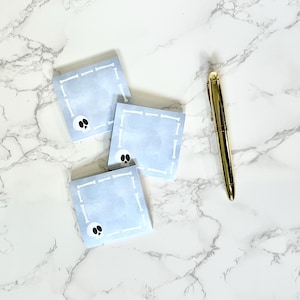May include: Three blue square sticky notes with a white bone border and a black skull graphic in the center. The notes are on a white marble surface with a gold pen.