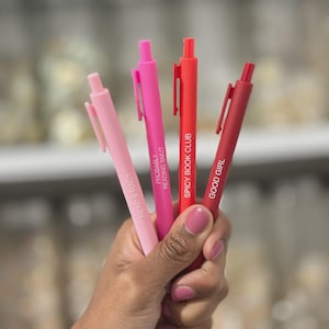 May include: A hand holds four pens in varying shades of pink and red. Each pen is inscribed with a different phrase: "READ & RACY", "PROBABLY READING SMUT", "SPICY BOOK CLUB", and "GOOD GIRL".