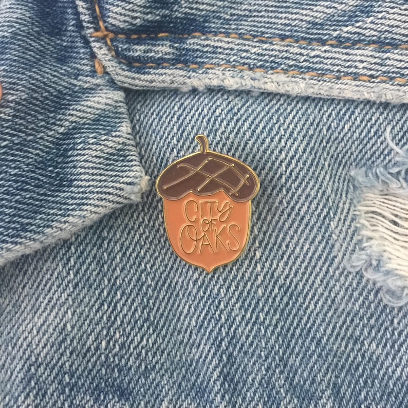 May include: Gold acorn-shaped pin with a brown top and a peach-colored bottom. The pin says "City of Oaks" in gold lettering.